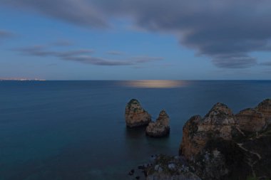 Bir deniz ay ışığının yüzey ve uzak şehre su ufuk üzerinde. Sessiz bir su yüzey ve kayalık uçurum geceleri Lagos, Portekiz.