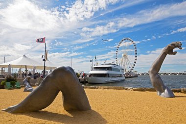 OXON HILL, MARYLAND, USA - SEPTEMBER 11, 2016: Awakening, a sculpture at National Harbor in front of pier and Ferris wheel. A famous statue of a giant embedded in the earth created by J. Seward Johnson Jr. clipart