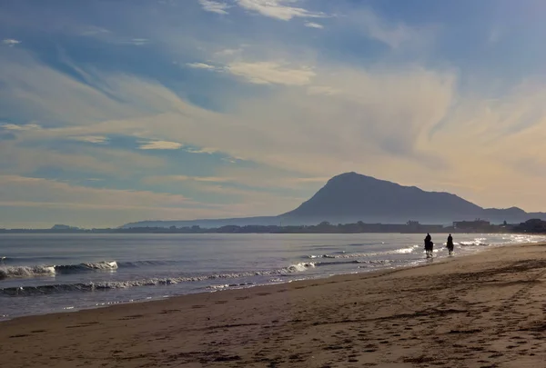 Waves roll on a sandy beach with horse riders. Clouds formation over mountains and the beach of Mediterranean Sea near Denia, Valencia, Spain.