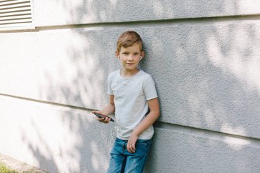 Uzağa arıyorsunuz genç çocuk kulaklık takıyor. 7 yaşında çocuk ile smartphone. Çocuk sarışın genç çocuk açık havada portresi. kulaklık ile müzik dinleme