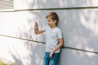 Uzağa arıyorsunuz genç çocuk kulaklık takıyor. 7 yaşında çocuk ile smartphone. Çocuk sarışın genç çocuk açık havada portresi. kulaklık ile müzik dinleme