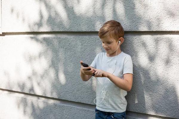Young boy wearing headphones, looking away. 7 years old boy with smartphone. Portrait of Child blond young boy outdoors. listening music  with headphones