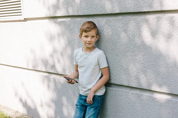 Uzağa arıyorsunuz genç çocuk kulaklık takıyor. 7 yaşında çocuk ile smartphone. Çocuk sarışın genç çocuk açık havada portresi. kulaklık ile müzik dinleme