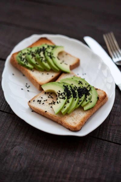 Delicious toast with avocado slices and black sesame. healthy breakfast concept. toast on white plate. making sandwiches with avocado on dark wooden background. healthy organic food top view.