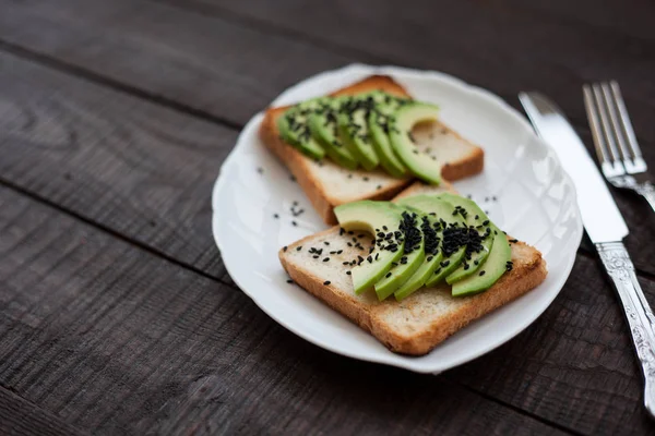 Delicious toast with avocado slices and black sesame. healthy breakfast concept. toast on white plate. making sandwiches with avocado on dark wooden background. healthy organic food top view