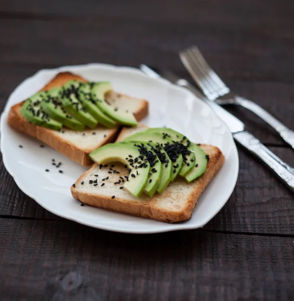 Delicious toast with avocado slices and black sesame. healthy breakfast concept. toast on white plate. making sandwiches with avocado on dark wooden background. healthy organic food top view.