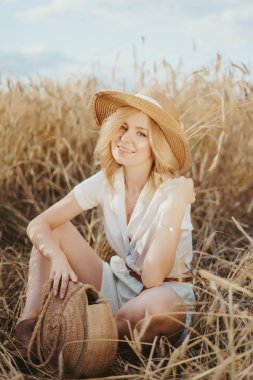 Sarı günbatımı tarlasında altın hasadında oturan mutlu genç kızın portresi. Yaz doğası geçmişi. Hasır şapkalı sarı saçlı kadın. Genç kadın buğday tarlasında oturuyor.