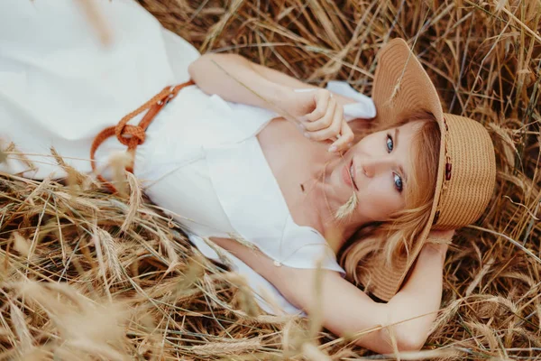 Beauty Girl Outdoors enjoying nature. Beautiful blonde Model girl in white dress on the Summer wheat Field, Sun Light. Girl Outdoors enjoying nature. Happy stylish woman. straw hat
