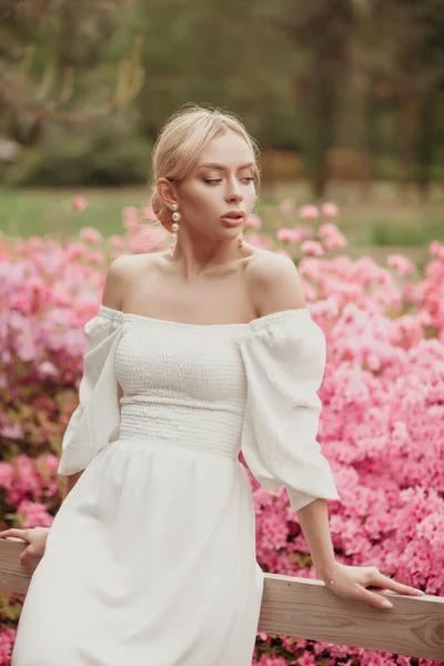 Genç ve güzel bir kadın parkta poz veriyor. Beyaz elbiseli bayan. Pembe çiçekli arka plan. Sarışın model. romantik bahar portresi