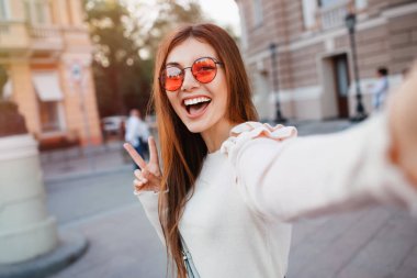 Selfie-sokak arka plan, uzun esmer saçlı güzel kız portresi. bej elbise, kırmızı güneş gözlüğü, Makyaj giyer. İki parmak gösterilen selfie, yapım gözlük gülümseyen ve positive.young kadın
