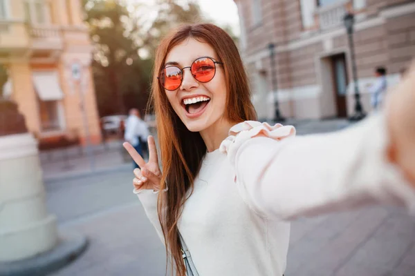 Selfie の通りの背景で長いブルネットの髪を持つ美少女の肖像画 ベージュのドレス 赤いサングラス メイクアップを着ています 本の指を示す Selfie を作るガラスの笑顔と Positive Young — ストック写真