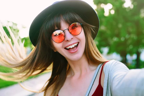 若い女の子ストリート夏市の携帯電話を持つ手から Selfie を取る 都市生活のコンセプトです スタイリッシュな帽子とドレスを身に着けている Selfie 笑みを浮かべて 面白い顔を作る長い髪の流行に敏感な若い女性 — ストック写真