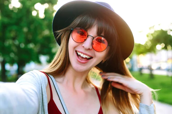 若い女の子ストリート夏市の携帯電話を持つ手から Selfie を取る 都市生活のコンセプトです スタイリッシュな帽子とドレスを身に着けている Selfie 笑みを浮かべて 面白い顔を作る長い髪の流行に敏感な若い女性 — ストック写真