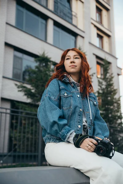 Bella ragazza dai capelli rossi in posa con una macchina fotografica sulla strada vicino a un edificio di mattoni in una serata estiva — Foto Stock