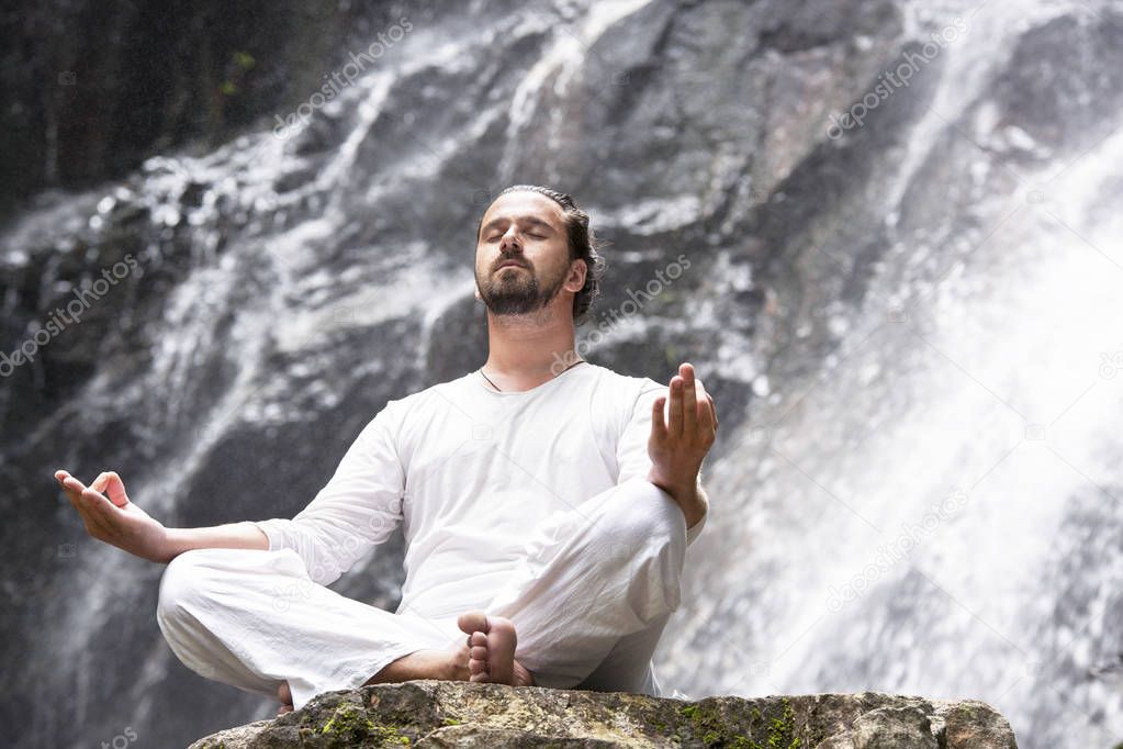 Wellness yoga meditation concept. Young man sitting in lotus position on the rock under tropical waterfall.