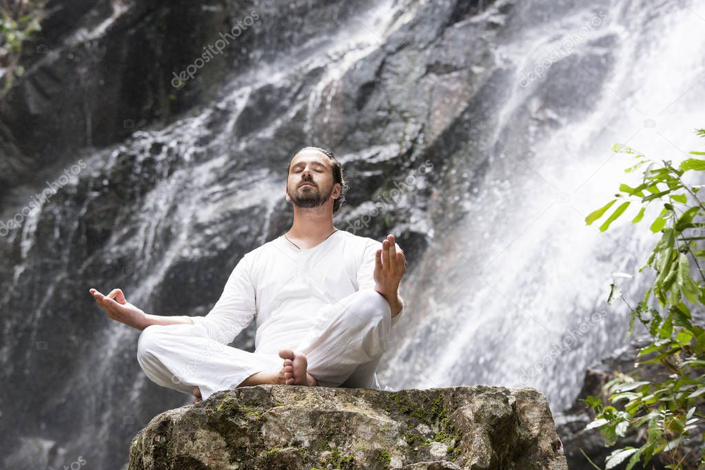Wellness yoga meditation concept. Young man sitting in lotus position on the rock under tropical waterfall.