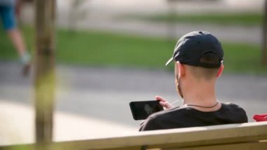 Genç adam parkta oturuyor ve akıllı telefondan sohbet ediyor. Mutlu adam akıllı telefon kullanıyor. Şehir parkında bankta oturuyor..