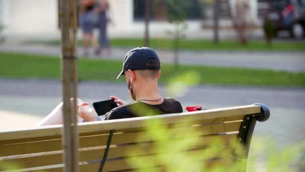 Молодой человек сидит в парке и болтает по смартфону. Счастливчик пользуется смартфоном, сидя на скамейке в городском парке. — стоковое видео