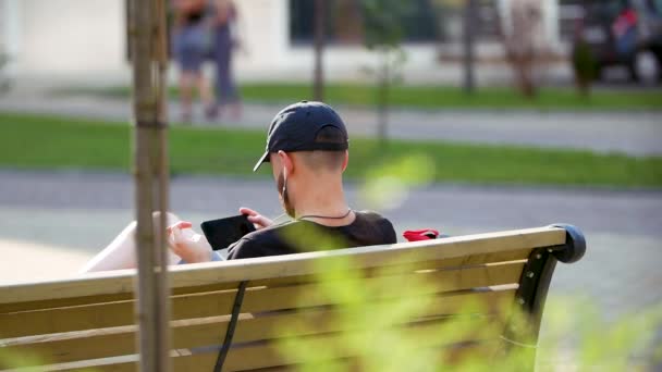 De jongeman zit in het park te kletsen op de smartphone. Happy man gebruik smartphone zitten op de bank in het stadspark. — Stockvideo