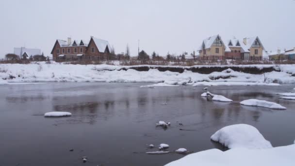 Paesaggio invernale - fiumi scongelati scorrono tra le ripide rive oltre cottage residenziali. Sta nevicando. . — Video Stock