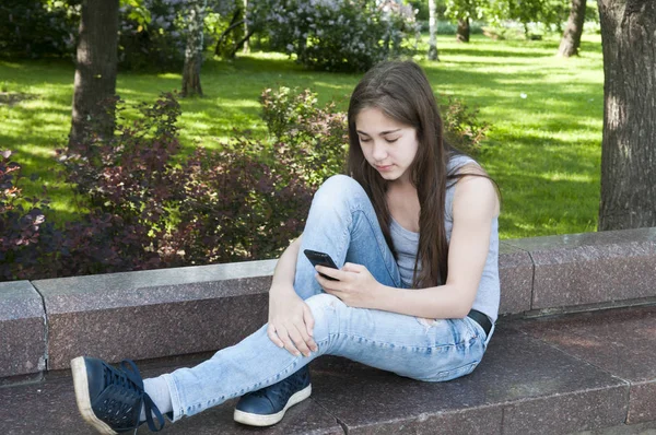 Молодая привлекательная девушка пользуется телефоном на скамейке. Летний парк. Фото — стоковое фото