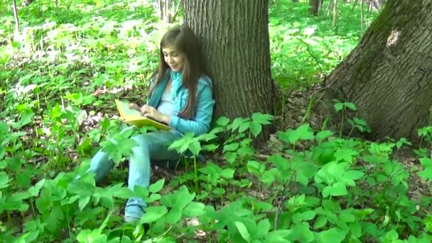 Açık eğitim genç kız kitap ve ormandaki ağaç gövdesi karşı ilkbaharda, eğilerek dönüm sayfa okuma. İlk ciddi, sonra güler. — Stok video