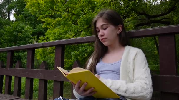 Retrato de una adolescente seria leyendo libro y volteando la página apoyada en el banco en el bosque en primavera, estudiando al aire libre. Vídeo HD filmación de la cámara estática . — Vídeos de Stock