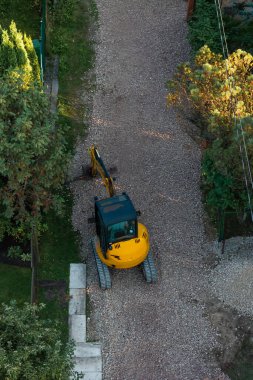A yellow excavator with a black cabin rests on a gravel pathway bordered by grass and concrete slabs, with power lines visible to the right.