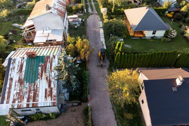 Aerial view of a residential area featuring a gravel pathway, a weathered building with a rusted roof, a modern house, and a small excavator at work.