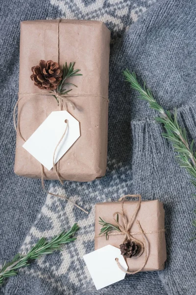 Christmas gifts packed with eco-friendly materials on the background of a gray knitted sweater with an ornament. Boxes wrapped in kraft paper, decorated with pine cones and rosemary branches.