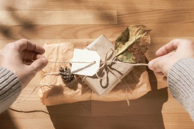 Gri kazak giymiş eller sonbahar tatili için hediye paketliyor. Şükran Günü, Cadılar Bayramı. Sahne, ahşap parke ve parlak güneş ışığından oluşan bir zemin üzerinde fotoğraflandı. Üst görünüm.