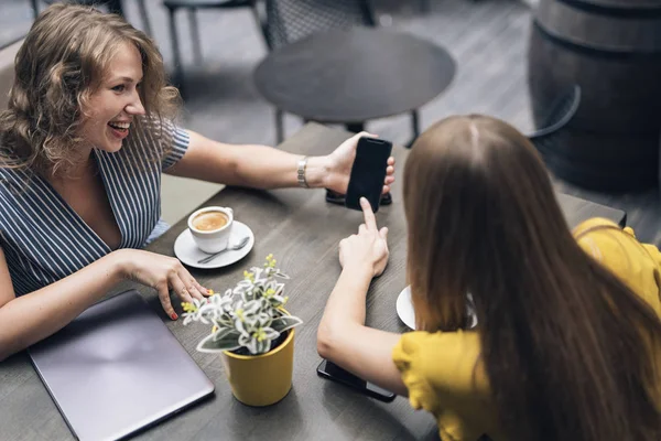 Contemporary female friends enjoying coffee at table in cafe while sharing smartphone and smiling