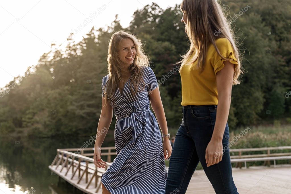 Contemporary young girlfriends walking on wooden pier of lake in sunlight and chatting