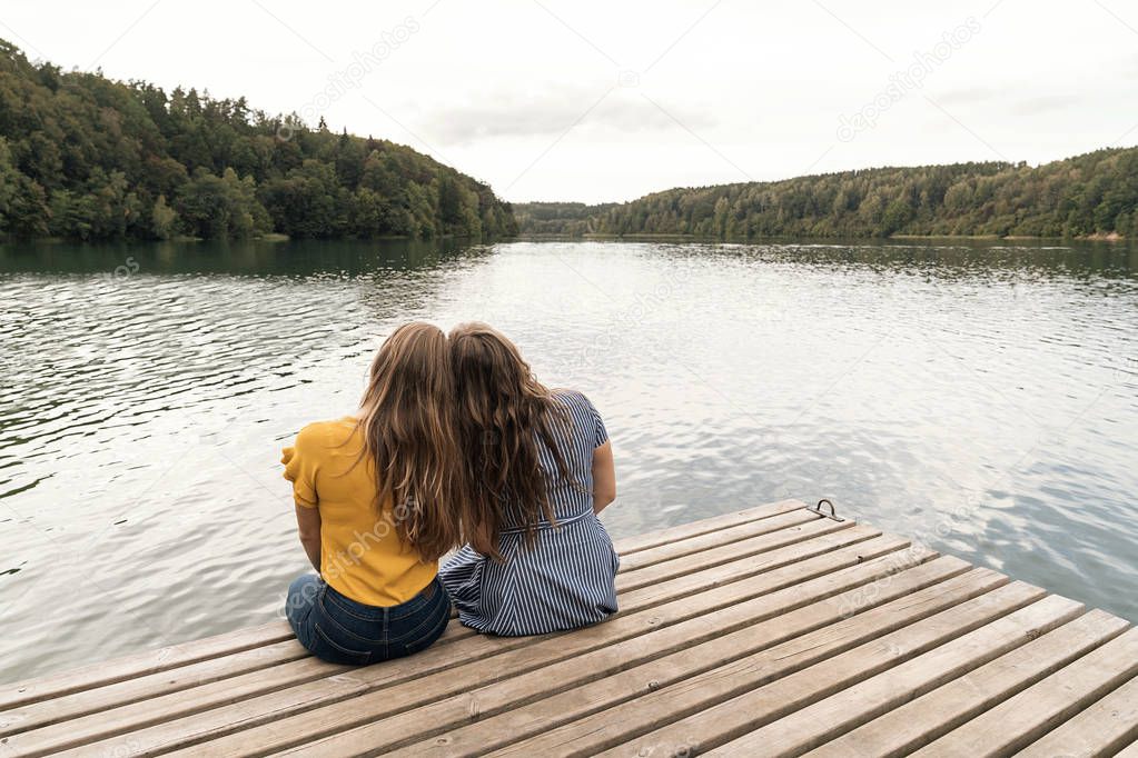 Back view of women sitting close on wooden pier against landscape of lake and forests