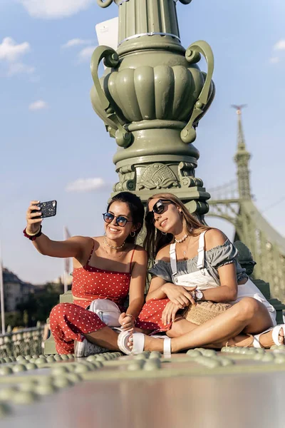 Mulheres alegres sentadas na ponte e usando celular — Fotografia de Stock