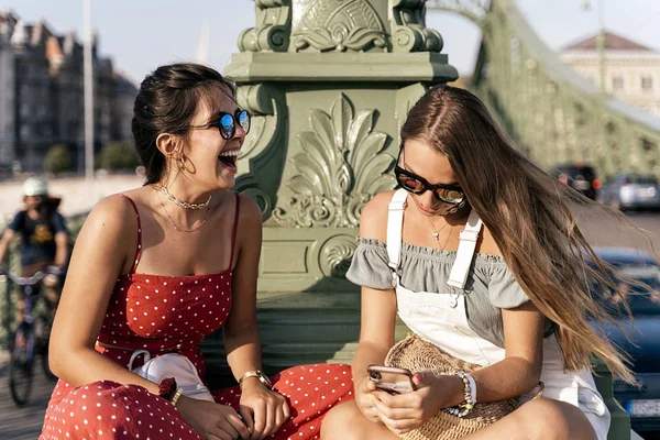 Cheerful women sitting on bridge and using cellphone — Stock Photo, Image