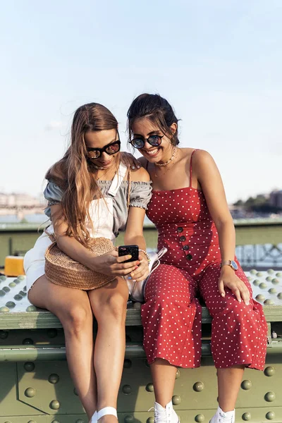 Happy female friends sitting on bridge and using smartphone — Stock Photo, Image