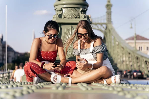 Wesołe kobiety siedzące na mostku i za pomocą telefonu komórkowego — Zdjęcie stockowe