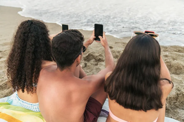 Amigos sentados con teléfonos inteligentes en la orilla del mar de arena — Foto de Stock