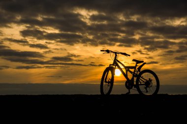 Bir bisiklet sahilde duruyor. Arka planda sarı bir gökyüzü günbatımı. Tüm gökyüzü küçük kara bulutlar ile kaplıdır. Sarı güneşin Bisiklet siluet siyah parlar.