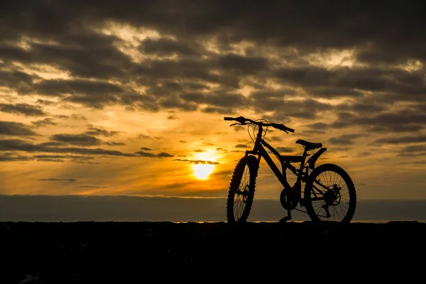 A bicycle stands on the beach. In the background a yellow sky at sunset. The whole sky is covered with small dark clouds. The sun is almost at the horizon in the center of the frame. To the right of the frame is a black silhouette of a bicycle.