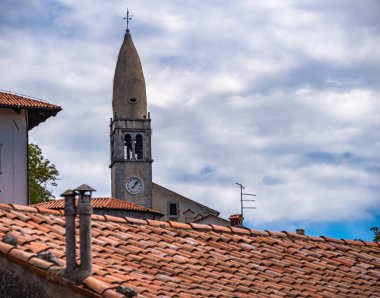 Stanjel, Slovenya - 27 Temmuz 2025: Saati olan bir kilise kulesi bulutlu bir gökyüzünün altında terasotta çatılarının üzerinde yükseliyor.