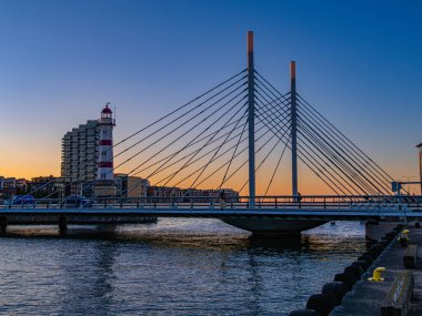 Malmö, İsveç - 6 Eylül 2025: Günbatımında nehrin üzerinde deniz feneri ve şehrin silueti olan modern bir köprü.