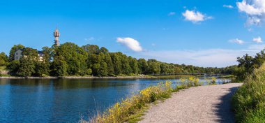 Stockholm, İsveç - 5 Eylül 2025: Ağaçlı nehir yolu ve mavi gökyüzünün altında bir kule.