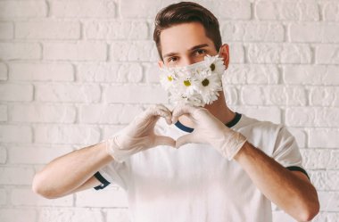 Tıbbi eldivenler ve çiçek koruyucu yüz maskesi takan genç adamın portresi kalp sembolünü elleriyle gösteriyor. Yaz sıhhiyesi maskeli mutlu erkek doktor parmaklarıyla aşk işareti şeklinde hareket ediyor. Hayır Kurumu, COVID-19