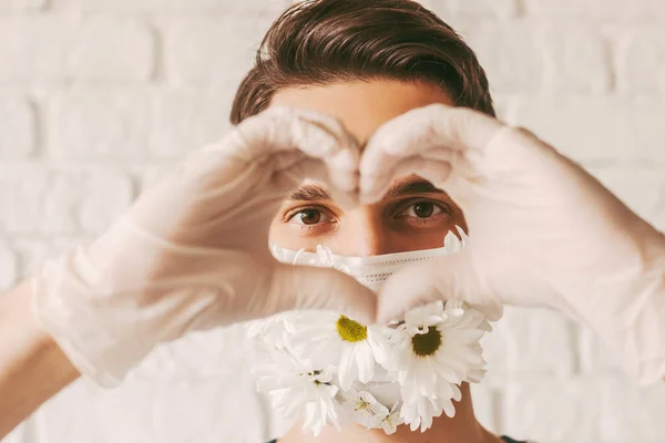 Portrait happy male doctor in medical face mask with flowers looking through hands in shape of heart symbol. Young man in protective face mask and medical gloves gesturing love sign. Charity, COVID-19