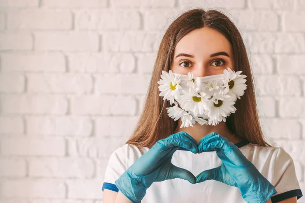 Yazın, tıbbi maske takan ve koruyucu eldiven takan mutlu genç bir kadının portresi kalp şeklini ve gülümsemesini gösteriyor. Çiçekli, koruyucu maskeli güzel bir doktor aşk sembolü. Güzel, hayır.