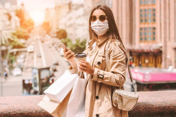 Happy elegant woman in protective face mask with shopping bags on sunny city street. Beautiful stylish hipster girl shopper in medical face mask drink coffee, use mobile phone after shopping in mall