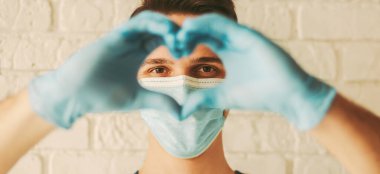 Tıbbi maske takan mutlu bir doktor ve kalp şeklini işaret eden koruyucu eldivenler. Koruyucu maskeli genç bir adam ve aşk sembolü gösteren tıbbi eldivenler. Sağlık, COVID-19