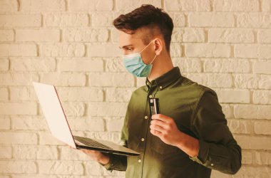 Tıbbi maskeli genç hippi elinde dizüstü bilgisayar ve kredi kartı tutuyordu. Çevrimiçi mağazadan koruyucu maskeli bir adam. Coronavirus COVID-19 karantinası. Erkek alışverişçi. İnternetten güvenli alışveriş